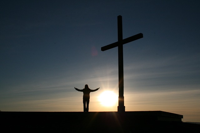 Sunrise silhouette of a person standing with arms out next to a cross.