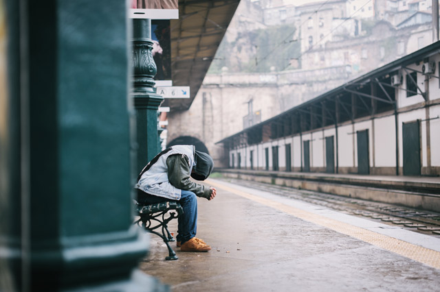 A man, bent over low, sitting at a railway station.