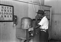 A mid-20th century photo showing a Black man drinking from a segregated drinking fountain