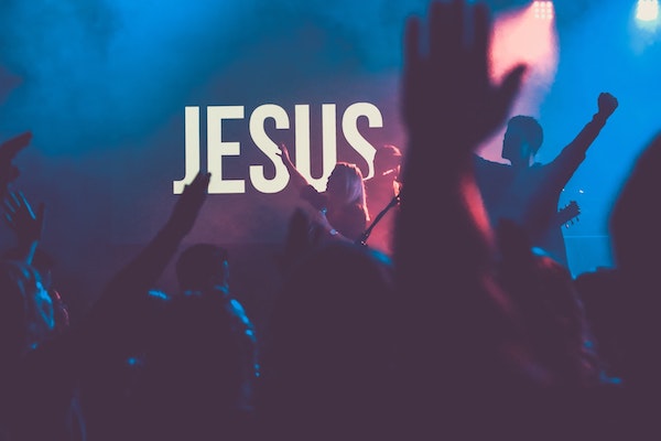 A crowd of people in a dark room in front of a sign that says "JESUS" with their hands raised in worship